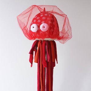 May include: A handmade jellyfish decoration with a red fabric head adorned with white floral patterns and large, googly eyes. A red mesh veil surrounds the head, and long, colourful fabric strips form the tentacles. Designed to hang, this is a unique piece.