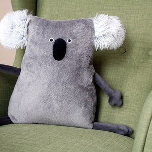 May include: A gray koala-shaped plush pillow with a black nose and eyes, and fluffy white ears. The stuffed animal is sitting on a green armchair. The koala has gray arms and legs.