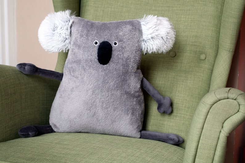 May include: A gray koala-shaped plush pillow with a black nose and eyes, and fluffy white ears. The stuffed animal is sitting on a green armchair. The koala has gray arms and legs.