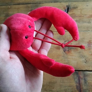 May include: A red plush lobster toy with black eyes. The lobster is curled up with its claws together.