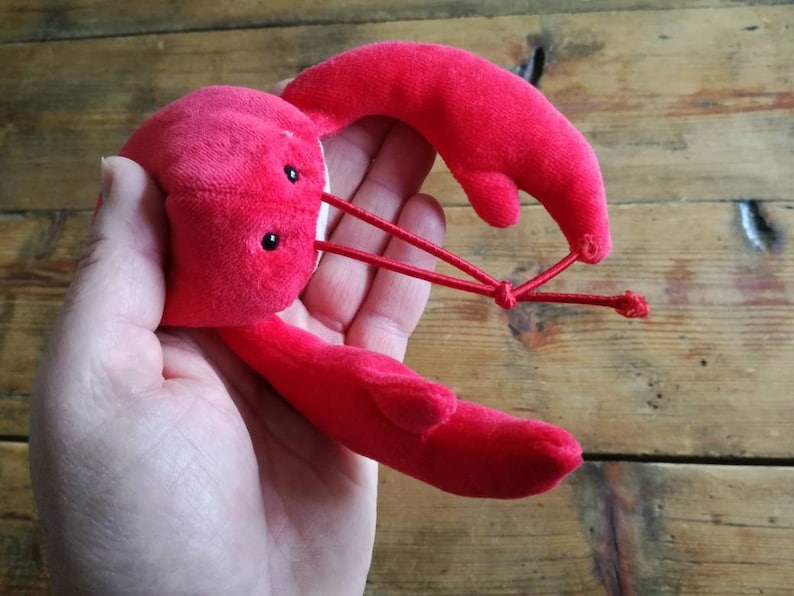 May include: A red plush lobster toy with black eyes. The lobster is curled up with its claws together.