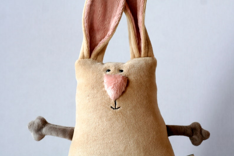 Pu&ograve; includere: Un peluche a forma di coniglio con corpo beige, interno orecchie rosa e naso rosa. Il coniglio ha occhi neri a bottone e dettagli ricamati. Le sue braccia sono di una tonalit&agrave; pi&ugrave; scura di marrone. Un giocattolo morbido per bambini.