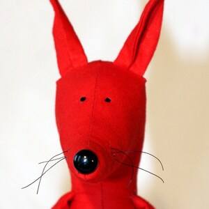 May include: A red plush toy with long ears, a black nose, and black whiskers. The toy is made of soft fabric and has a simple design.