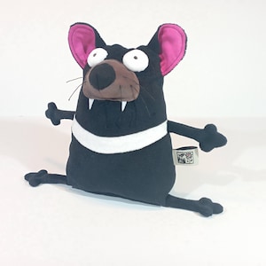 May include: A plush Tasmanian devil toy in black with a white stripe, pink-lined ears, and a brown snout. The toy has large white eyes, visible fangs, and outstretched arms and legs. A small tag is attached to the toy.