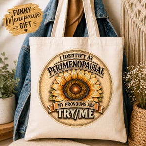 May include: Cream-colored tote bag with a sunflower design and the text "I Identify as Perimenopausal. My Pronouns Are Try/Me." A small, brown, oval-shaped graphic reads "Funny Menopause Gift". The bag has two handles.