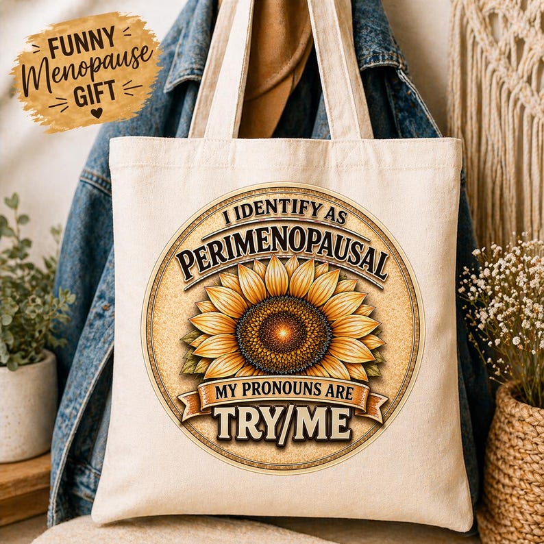 May include: Cream-colored tote bag with a sunflower design and the text "I Identify as Perimenopausal. My Pronouns Are Try/Me." A small, brown, oval-shaped graphic reads "Funny Menopause Gift". The bag has two handles.