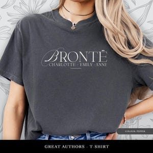 Puede incluir: Camiseta gris oscuro con la palabra "BRONTE" en elegante escritura blanca, con "CHARLOTTE - EMILY - ANNE" debajo. La camiseta es de la colección "GREAT AUTHORS - T-SHIRT" y es de color pimienta.
