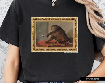 Pode incluir: T-shirt preta com uma pintura emoldurada de um gato em uma moldura dourada. A pintura retrata um gato batendo em objetos em uma mesa. O texto "ART HISTORY SERIES - T-SHIRT" está na parte inferior.