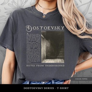 May include: Pepper-colored t-shirt featuring the title "DOSTOEVSKY" in an elegant font above a vintage-style image and text from "Notes From Underground." The shirt's design includes a faded, distressed look, with the text "DOSTOEVSKY SERIES - T-SHIRT" at the bottom.