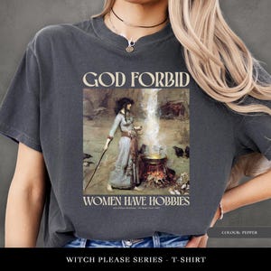 May include: Pepper-coloured t-shirt with a vintage illustration and the text "GOD FORBID WOMEN HAVE HOBBIES." The graphic shows a woman in a long dress with a staff, near a cauldron. Part of the "Witch Please Series" range.
