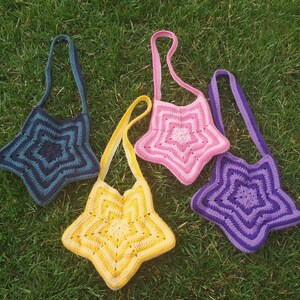 May include: Four handmade crochet star-shaped bags in shades of blue, pink, yellow, and purple. Each bag has a long strap for over-the-shoulder wear. The bags are displayed on a bed of green grass, showcasing their vibrant colors and unique design.