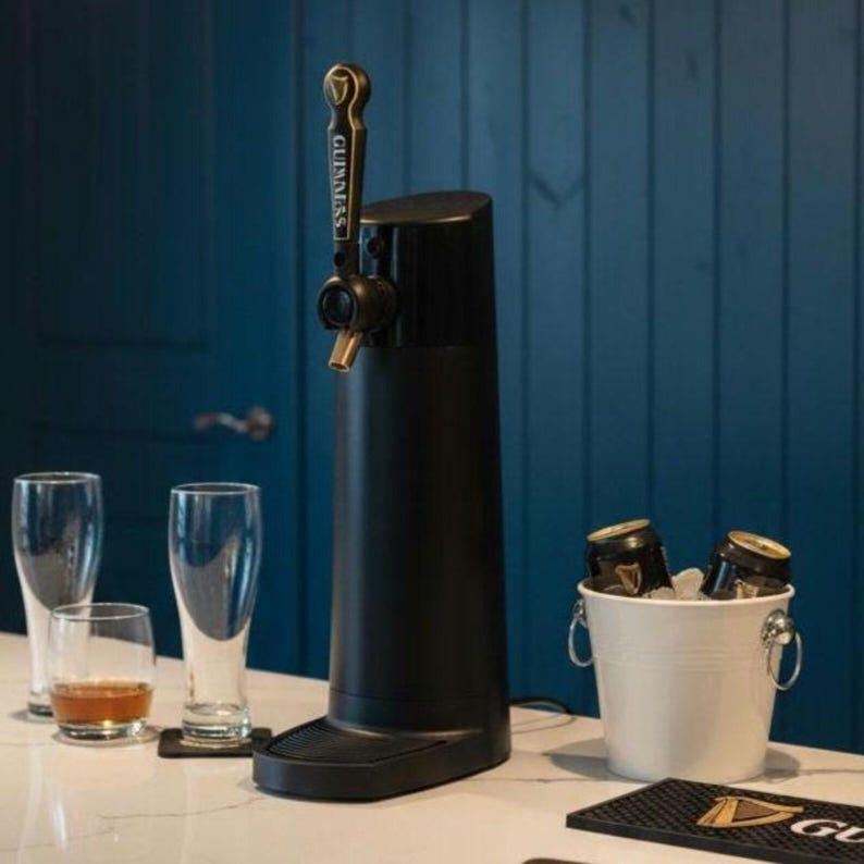 May include: A black beer tap dispenser with a gold handle, next to a white bucket with ice and beer cans. There are also beer glasses and a small glass with amber liquid on a white countertop. The background is a blue wall.