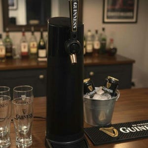 Könnte beinhalten: Ein schwarzes Guinness-Zapfsystem mit einem Zapfhahn mit dem Guinness-Logo. Zwei Pintgläser und ein kleiner Eimer mit Eis und Guinness-Dosen stehen auf der Holzbar. Eine schwarze Barmatte mit dem Guinness-Logo ist ebenfalls sichtbar.