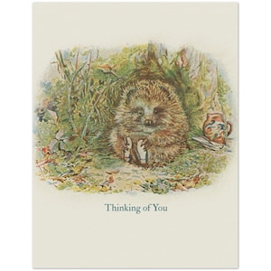 May include: A greeting card featuring a watercolor illustration of a brown hedgehog sitting in a green, leafy environment. Beside the hedgehog is a small, patterned ceramic pitcher. The text "Thinking of You" is printed in blue at the bottom.