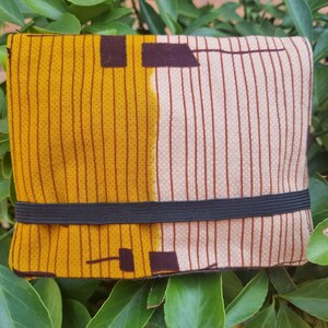 Può includere: Un portafoglio rettangolare in tessuto con un motivo sorprendente. Il design presenta una combinazione di pannelli giallo senape e beige, ciascuno con strisce verticali. Accenti marrone scuro e un elastico nero completano il design.