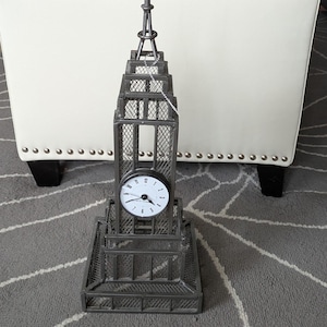 May include: A decorative metal clock shaped like the Empire State Building. The clock has a round face with black numbers and hands. The building is gray with a detailed cage-like design and a pointed spire.