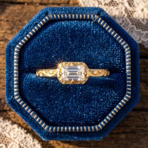 Könnte beinhalten: Ein goldener Ring mit einem rechteckigen, im Smaragdschliff geschliffenen, klaren Edelstein in einer texturierten Fassung. Die Ringschiene zeigt beidseitig eingravierte Rankenmuster und liegt in einer achteckigen, dunkelblauen Samtschatulle.