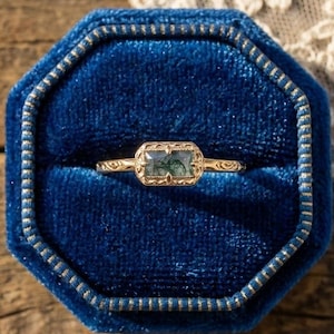 Op de afbeelding: Een gouden ring met een rechthoekige, mosgroene edelsteen in een decoratieve gouden band. De ring wordt gepresenteerd in een blauwe fluwelen achthoekige doos, tegen een houten oppervlak.