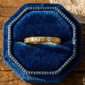 May include: A gold ring with a repeating pattern of starbursts and small diamonds, set in a blue velvet ring box. The ring is displayed on a wooden surface, with a lace fabric in the background.