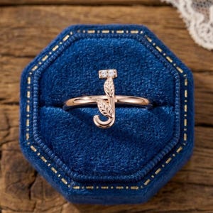 May include: A rose gold ring with a leaf-shaped design and a small gemstone at the top. The ring is in the shape of the letter "J" and is displayed in a blue velvet ring box.