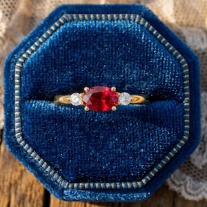 Peut inclure: Une bague en or avec une pierre centrale ovale rouge et deux petits diamants de chaque côté, présentée dans un écrin en velours bleu. La bague est posée sur une surface en bois avec de la dentelle.