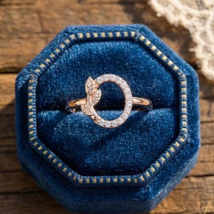 Op de afbeelding: Een roségouden ring met een bladmotief en een met diamanten bezette letter "O", gepresenteerd in een blauwe fluwelen ringdoos. De ring wordt getoond op een houten oppervlak, met kant op de achtergrond.