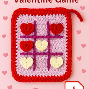以下が含まれることがあります： 赤、白、紫のアクセントが施された手作りのかぎ針編みの三目並べゲーム。ゲーム盤は正方形で赤い縁取りがあり、ハート型のゲームピースが特徴です。「Handmade Couple Valentine Game」というテキストが上部にあります。