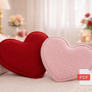 May include: Two heart-shaped pillows, one red and one pink, rest on a white bed. The pillows are made of a textured material, likely knitted or crocheted. The background is softly lit, suggesting a cozy, romantic setting. A PDF icon is visible.