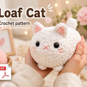 Könnte beinhalten: Ein weißes Häkeltier in Katzenform mit rosa Akzenten an Ohren und Gesicht, gehalten in Händen. Die Katze hat schwarze Augen und einen gestickten rosa Mund. Der Text oben lautet "Loaf Cat Crochet pattern". Ein PDF-Symbol befindet sich unten links.