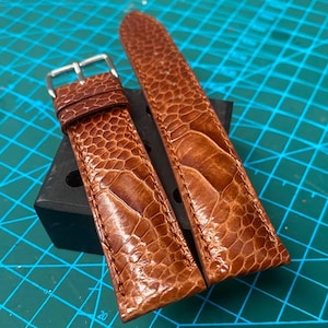 Puede incluir: Dos correas de reloj de cuero marrón con un patrón texturizado y una hebilla plateada. Las correas se muestran sobre un bloque negro contra un fondo de cuadrícula turquesa. El cuero tiene un aspecto de reptil.