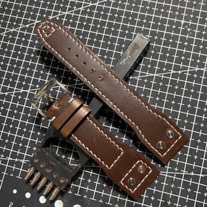 Puede incluir: Dos correas de reloj de cuero marrón con costuras blancas y herrajes plateados se muestran sobre una esterilla de corte negra. Las correas tienen un diseño clásico, con hebilla y remaches. También se ve una herramienta de metal.