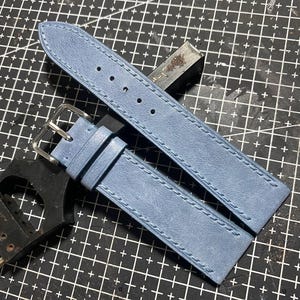 May include: A light blue watch strap with a silver buckle and white stitching. The strap is made of a textured material and is shown on a black cutting mat with other tools.
