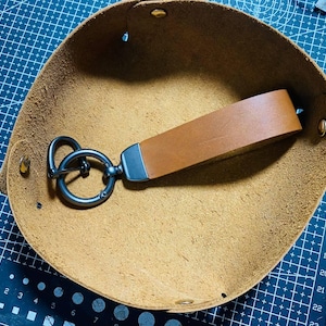 May include: A brown leather keychain with a silver-tone metal ring and clasp rests inside a round, tan leather tray. The tray has a textured surface and is secured with rivets. The background features a blue cutting mat with measurement markings.