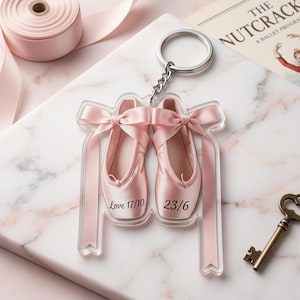 May include: A clear acrylic keychain featuring two pink ballet slippers with ribbons and bows. The keychain has a silver ring and chain. The text "Love 17/10" and "23/6" is printed on the slippers. A roll of pink ribbon is in the background.