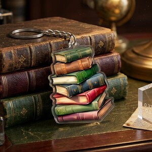 May include: A keychain featuring a stack of colorful books. The books are in shades of green, brown, red, and blue. The keychain is attached to a silver chain and ring. A clear acrylic block with measurements of 0.32cm and 0.13in is also visible.