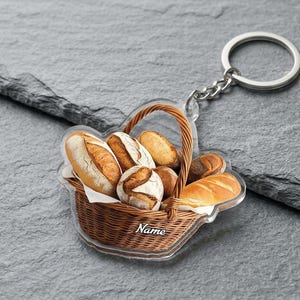 May include: A clear acrylic keychain featuring a basket of various breads. The basket is brown with a handle and contains several loaves of bread. The keychain has a silver-colored ring and chain. The word "Name" is written on the basket.