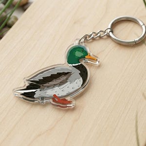 May include: A clear acrylic keyring with a detailed illustration of a mallard duck. The duck has a green head, yellow beak, and grey and black body. The keyring includes a silver chain and ring.