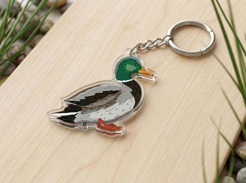 May include: A clear acrylic keyring with a detailed illustration of a mallard duck. The duck has a green head, yellow beak, and grey and black body. The keyring includes a silver chain and ring.