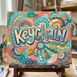 May include: A wooden artwork with the word "Keychain" painted in bold letters. The painting showcases a keychain with charms, including a cactus, star, cat, and tassel. The background is a mix of teal, orange, and white brushstrokes.