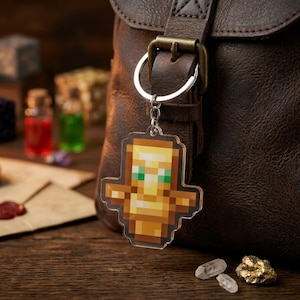 May include: A pixelated gold-colored keychain with a figure design and green eyes hangs from a silver ring. The keychain is in front of a brown leather bag. Other items on a wooden surface include a compass, potions, and crystals.