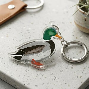 May include: A clear acrylic keyring with a detailed illustration of a mallard duck. The duck has a green head, grey body, and orange beak and feet. A silver keyring and a brown leather strap are also visible.