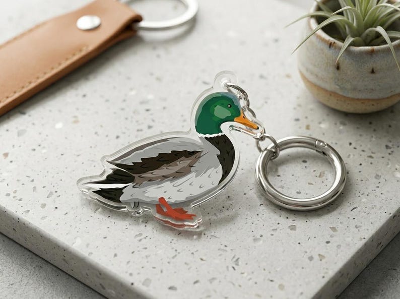 May include: A clear acrylic keyring with a detailed illustration of a mallard duck. The duck has a green head, grey body, and orange beak and feet. A silver keyring and a brown leather strap are also visible.