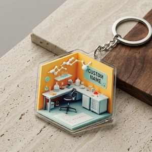 May include: A clear, hexagonal keychain featuring a miniature laboratory scene with a desk, lab equipment, and the text "CUSTOM NAME" on a light blue sign. The keychain has a silver keyring and chain.