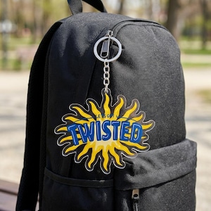 May include: A black backpack with a silver metal keychain attached to the zipper. The keychain features a yellow sun-shaped acrylic charm with the text "LET'S GET TWISTED" printed in blue letters across the center of the sun design.
