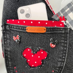 May include: Black denim bag with a red polka dot interior and a red Mickey Mouse applique. The bag features a pocket with a leather tag that reads "Hand Made". A smartphone is visible inside the bag.