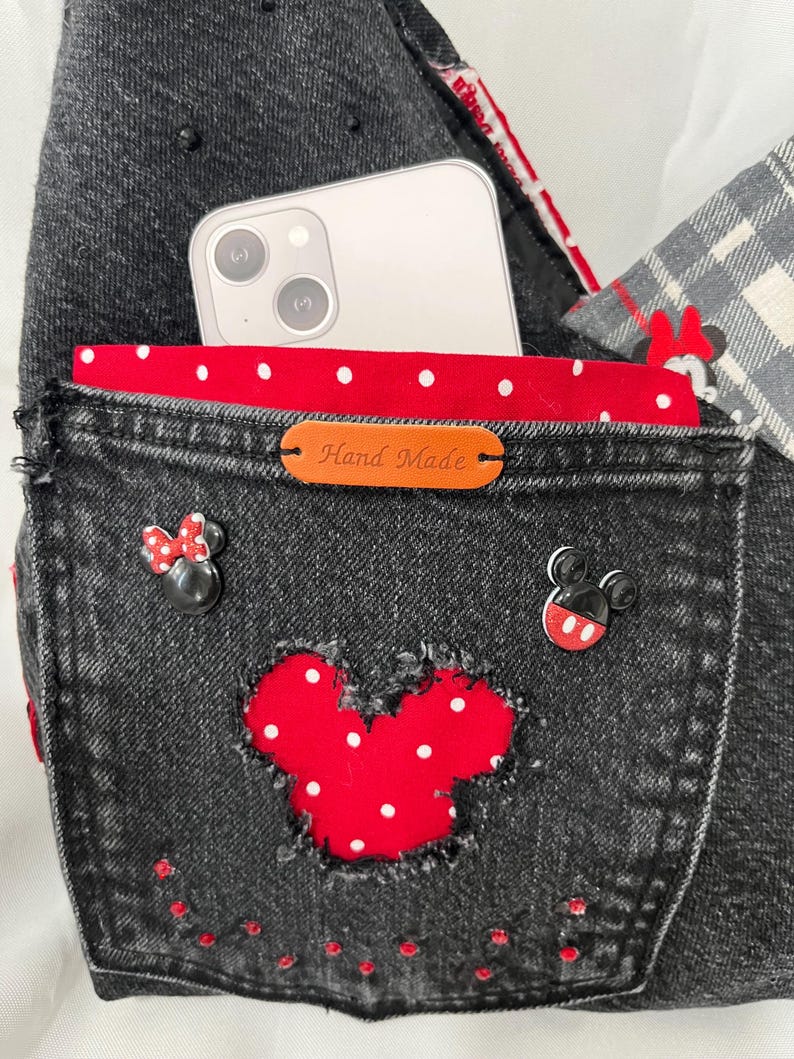 May include: Black denim bag with a red polka dot interior and a red Mickey Mouse applique. The bag features a pocket with a leather tag that reads "Hand Made". A smartphone is visible inside the bag.