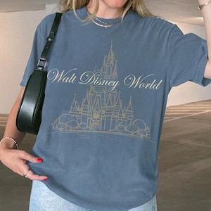 K&ouml;nnte beinhalten: Ein blaues T-Shirt mit einer goldenen Illustration eines Schlosses und den Worten "Walt Disney World". Das T-Shirt hat kurze &Auml;rmel und einen Rundhalsausschnitt. Das Design ist auf der Vorderseite des T-Shirts zentriert.