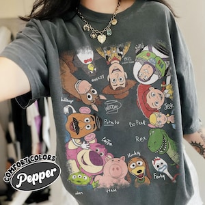 May include: Dark grey Comfort Colors t-shirt with a colourful graphic of Toy Story characters, including Woody, Buzz Lightyear, and Jessie. The shirt has the text "Pepper" and "COMFORT COLORS" on the left side.