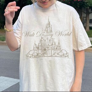 K&ouml;nnte beinhalten: Cremefarbenes T-Shirt mit einer detaillierten Illustration eines Schlosses und dem Text "Walt Disney World" in einer Schreibschrift. Das Design ist in ged&auml;mpften, neutralen T&ouml;nen gehalten und erzeugt eine Vintage-&Auml;sthetik. Das Shirt hat kurze &Auml;rmel und eine lockere Passform.