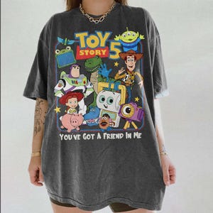 Pu&ograve; includere: T-shirt grigio scuro con una grafica colorata dei personaggi di "Toy Story 5". Il design include Buzz Lightyear, Woody e altri personaggi, con la scritta "YOU'VE GOT A FRIEND IN ME".